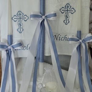 May include: A baptism set featuring white candles adorned with blue and white ribbons. The set includes a decorative bottle, a bar of soap, and a white towel embroidered with a cross design and the name "Nicholas".
