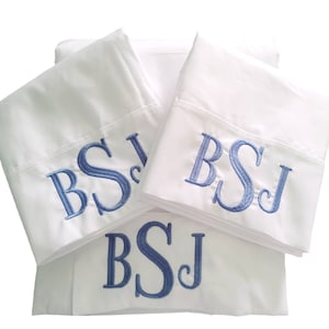 Pode incluir: Três fronhas brancas com iniciais bordadas em azul "BSJ" em cada uma. As fronhas estão empilhadas uma em cima da outra.