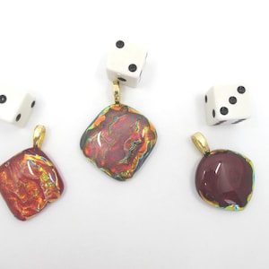 May include: Three square-shaped glass pendants with gold-toned bails, featuring iridescent red, orange, and dark maroon swirling patterns, accompanied by two white six-sided dice with black pips arranged in a scattered formation on a white surface.