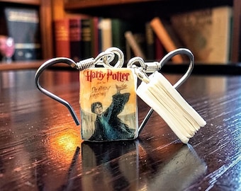 May include: A pair of silver-toned metal hook earrings featuring miniature book charms. One charm displays the Harry Potter and the Deathly Hallows cover art with a figure in a dark robe, while the other charm shows the white pages of a book.