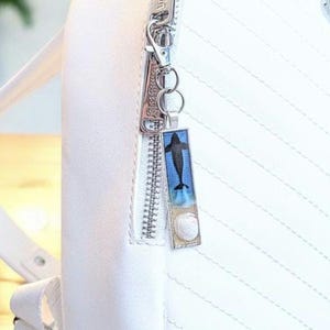 以下が含まれることがあります： 銀色のジッパーにぶら下がっているクジラのデザインの長方形のチャーム。チャームは、貝殻と砂の詳細を備えた青い背景に黒いクジラを特徴としています。チャームは白いバックパックに取り付けられています。