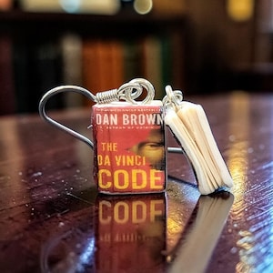 May include: A pair of miniature book earrings featuring a replica of The Da Vinci Code by Dan Brown. The front cover is red with yellow text and a partial face graphic. The earrings include silver-toned metal hooks and show white pages.