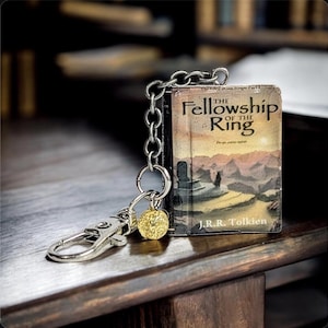 Pu&ograve; includere: Un portachiavi con un libro in miniatura intitolato "The Fellowship of the Ring" di J.R.R. Tolkien. Il libro ha un'illustrazione di copertina dettagliata ed &egrave; attaccato a una catena d'argento con un fermaglio e un ciondolo dorato.