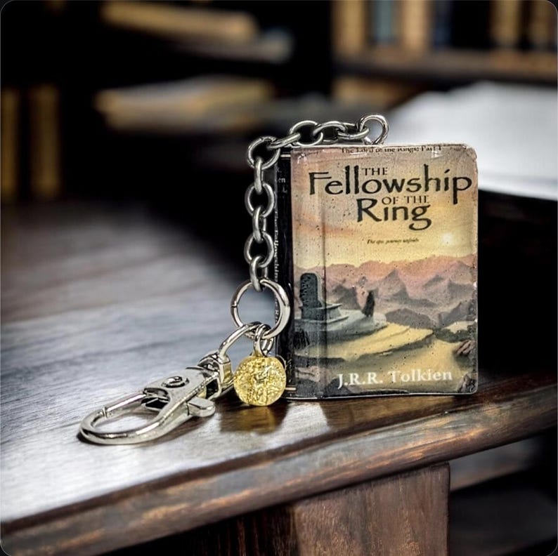 Pu&ograve; includere: Un portachiavi con un libro in miniatura intitolato "The Fellowship of the Ring" di J.R.R. Tolkien. Il libro ha un'illustrazione di copertina dettagliata ed &egrave; attaccato a una catena d'argento con un fermaglio e un ciondolo dorato.