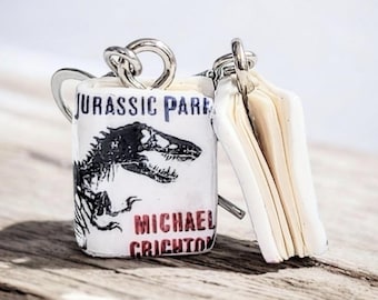 May include: A Jurassic Park-themed keyring featuring a small book and a cylindrical charm. The charm displays the book title, a dinosaur graphic, and the author's name in black and red text. The keyring is made of metal and white material.