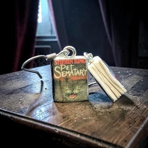 Peut inclure: Une paire de boucles d'oreilles pendantes avec des breloques en forme de livres miniatures. Chaque breloque est une réplique du roman Pet Sematary de Stephen King, avec le titre et l'auteur visibles sur la couverture, et des crochets en métal argenté.