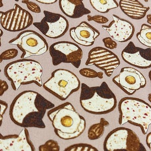 May include: A pink fabric with a repeating pattern of brown and white cats, toast, and fried eggs. The cats have brown and white fur and are depicted with their ears up. The toast is brown and white and has different toppings, including butter, jam, and sprinkles. The fried eggs are yellow with orange yolks.
