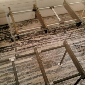 May include: A modern coffee table with a clear glass top and a metal frame. The frame is constructed with silver-colored pipes and wooden supports. The table is placed on a patterned rug.