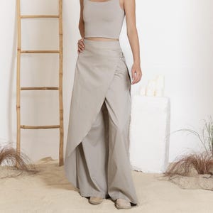 May include: A woman wearing a two-piece outfit in a neutral beige color. The top is a square-neck crop top, and the bottom is a wrap-style skirt over wide-leg pants. A bamboo ladder and decorative plants are in the background.