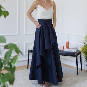 May include: A woman's outfit featuring a white cowl-neck camisole and a high-waisted, navy blue maxi skirt with a ruffled design. The skirt has a cinched waist and a flowing silhouette. The outfit is suitable for formal occasions.