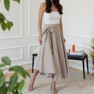 May include: A woman wearing a white tube top and a beige wrap skirt with a bow. The skirt falls to mid-calf length. She is wearing beige heels. The background is a white room with a black table and flowers.