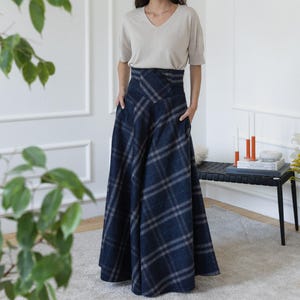 Könnte beinhalten: Eine Frau trägt ein beiges V-Ausschnitt-Oberteil und einen langen, marineblauen karierten Rock mit Taschen. Der Rock hat eine hohe Taille und eine ausgestellte Silhouette. Das Outfit befindet sich in einem hellen Raum.