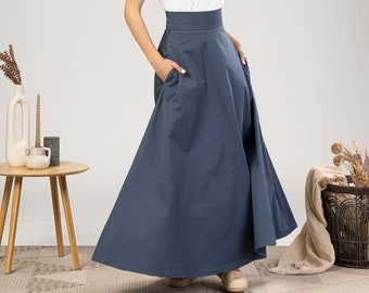 May include: A woman wearing a white short-sleeve t-shirt and a long, navy blue skirt with pockets. The skirt has a high waistband. The woman is smiling and has her hands in the skirt pockets. The skirt is floor-length.