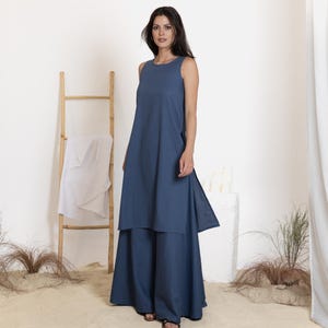 May include: A sleeveless, denim blue outfit consisting of a long tunic over wide-leg pants. The tunic has a high neckline and side slits. The pants are floor-length. The outfit is worn with black sandals. A wooden ladder and white fabric are in the background.