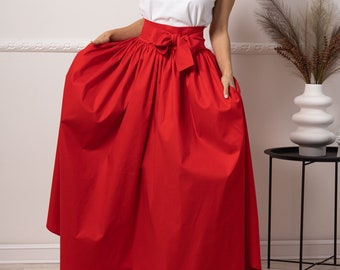 May include: A woman wearing a white camisole top and a long, vibrant red skirt with a bow at the waist. The skirt is floor-length and has a full, flowing design. The outfit is simple yet elegant.