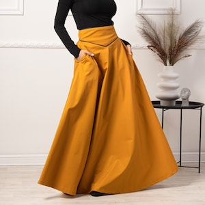 May include: A woman wearing a black long-sleeved turtleneck top and a mustard-yellow maxi skirt with pockets. The skirt has a black trim and a full, flowing silhouette. The outfit is set against a white wall.