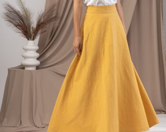 May include: A woman wearing a white camisole top and a long, mustard-yellow skirt. The skirt has a wide waistband and a flowing A-line shape. The background includes a neutral-toned curtain and a decorative vase.