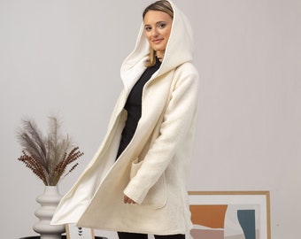 May include: A cream-colored hooded coat with a white interior and two front pockets. The coat is knee-length and has a relaxed fit. The model is wearing black leggings and boots. The coat is made of a soft, textured material.