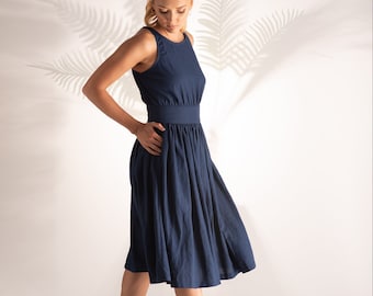 May include: A navy blue sleeveless dress with a fitted bodice and a flared skirt. The dress has a defined waist and falls to the mid-calf. The model is wearing black espadrille sandals with ankle ties. The background is a plain white wall with palm leaf shadows.