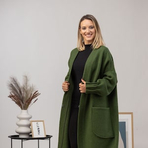 May include: An olive green long coat with large front pockets. The coat is open in the front and has a relaxed fit. The sleeves are long and the fabric appears to be a soft wool blend. The person is wearing a black top and black pants.