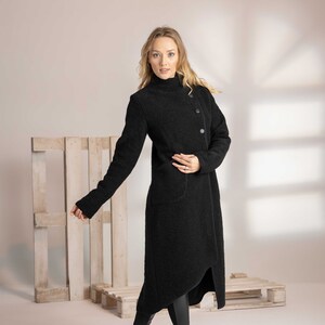 May include: A long black coat with a high collar and asymmetrical hemline. The coat features three large buttons and two pockets. The model is wearing black boots and leggings. The coat is made of a textured fabric.