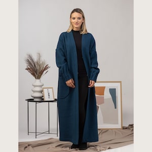 Könnte beinhalten: Langer, teal-farbener Cardigan mit lockerer Passform. Der Cardigan hat ein kragenloses Design, lange Ärmel und zwei große Vordertaschen. Das Model trägt ein schwarzes Oberteil und eine Hose. Der Cardigan ist bodenlang.