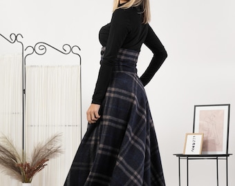 May include: A long, flared skirt with a navy blue and gray plaid pattern. The skirt is paired with a black long-sleeved top. The outfit is completed with a matching plaid belt. The model is smiling and looking over their shoulder.