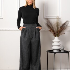 May include: A woman wearing a black turtleneck long-sleeve shirt and gray wide-leg trousers. The pants have a high waist and pockets. A white vase with decorative plants sits on a black side table.
