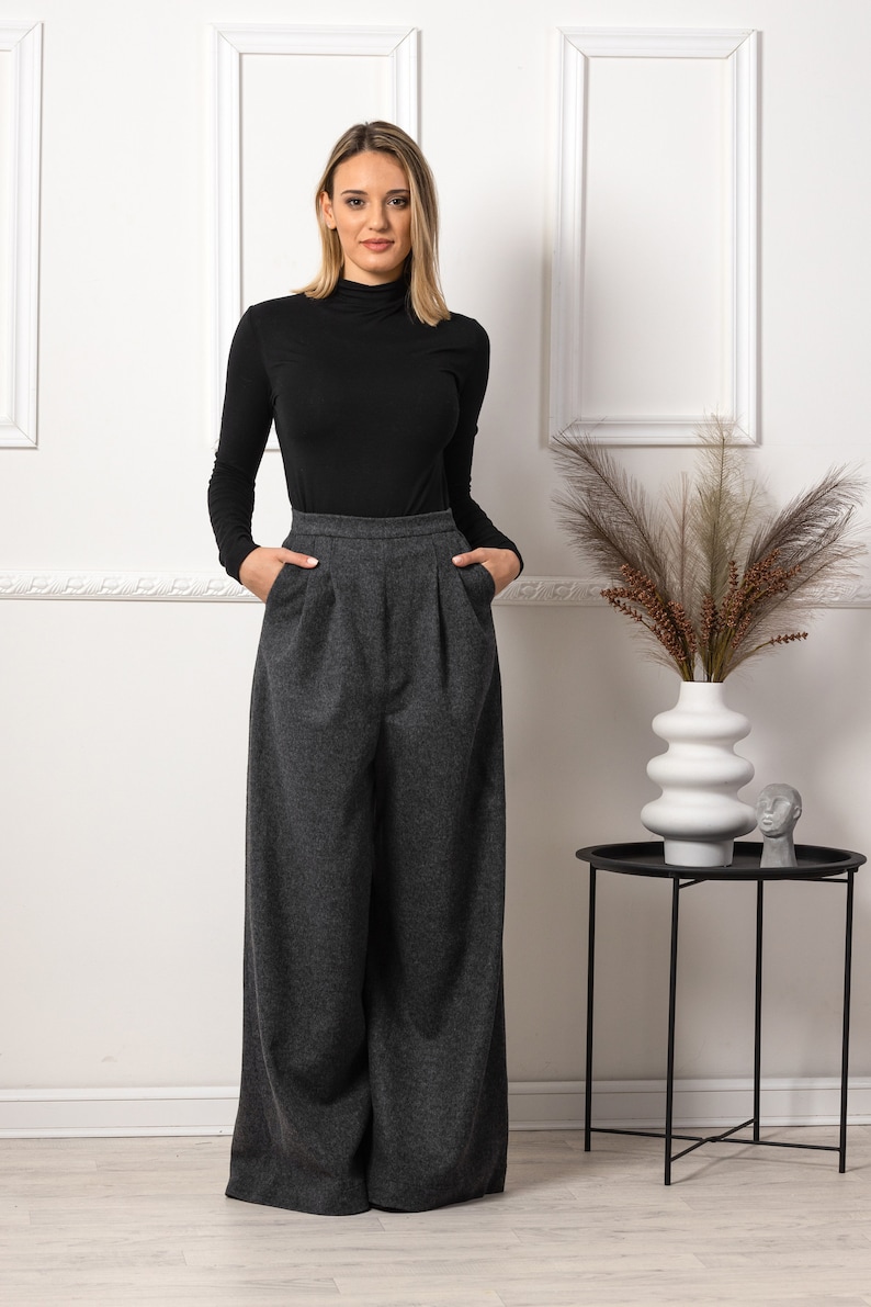 May include: A woman wearing a black turtleneck long-sleeve shirt and gray wide-leg trousers. The pants have a high waist and pockets. A white vase with decorative plants sits on a black side table.