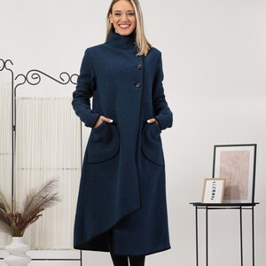 May include: A long, navy blue coat with a high collar and asymmetrical front. The coat features large pockets and black trim. It is buttoned with three black buttons. The model is wearing black boots and black leggings.
