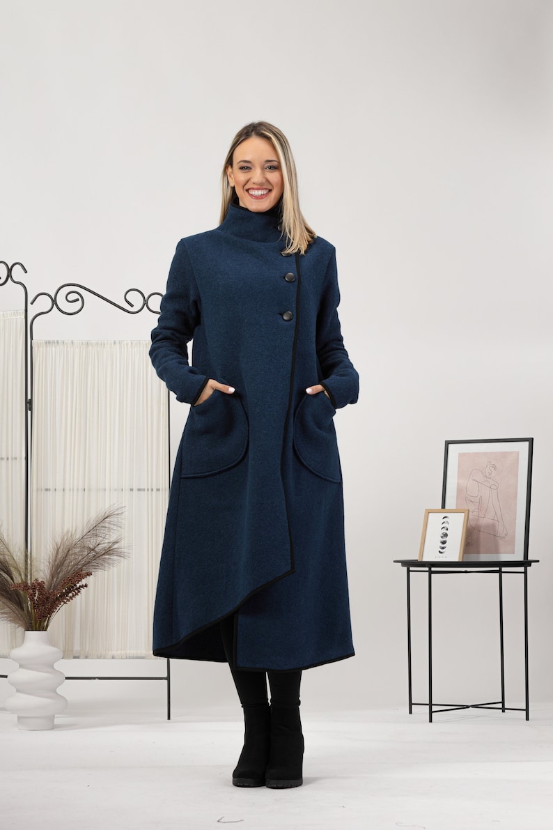 May include: A long, navy blue coat with a high collar and asymmetrical front. The coat features large pockets and black trim. It is buttoned with three black buttons. The model is wearing black boots and black leggings.