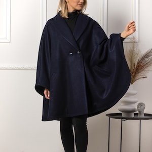 May include: A navy blue cape coat with a double-breasted front and large lapels. The coat has two front pockets and is worn with black leggings and boots. The coat is a stylish outerwear garment.