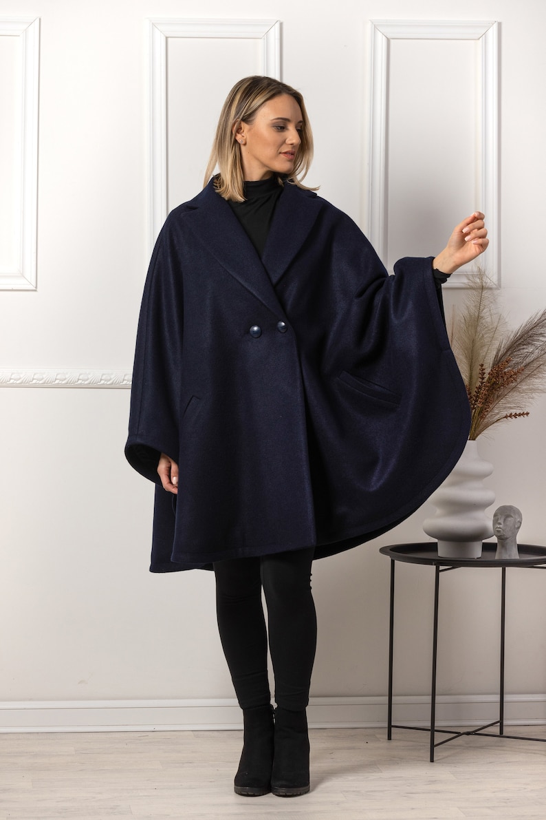 May include: A navy blue cape coat with a double-breasted front and large lapels. The coat has two front pockets and is worn with black leggings and boots. The coat is a stylish outerwear garment.