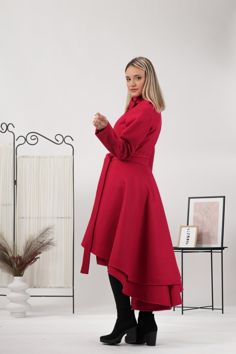 May include: A vibrant red coat featuring a high-low hemline, long sleeves, and a matching fabric belt tied at the waist. The coat has a structured collar and a flared silhouette that extends longer in the back, paired with black leggings and boots.