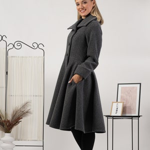 May include: A charcoal grey wool-blend coat featuring a wide collar, long sleeves with cuffs, a fitted bodice, and a flared, knee-length skirt with side pockets. The coat is fastened with a vertical row of buttons down the front centre.
