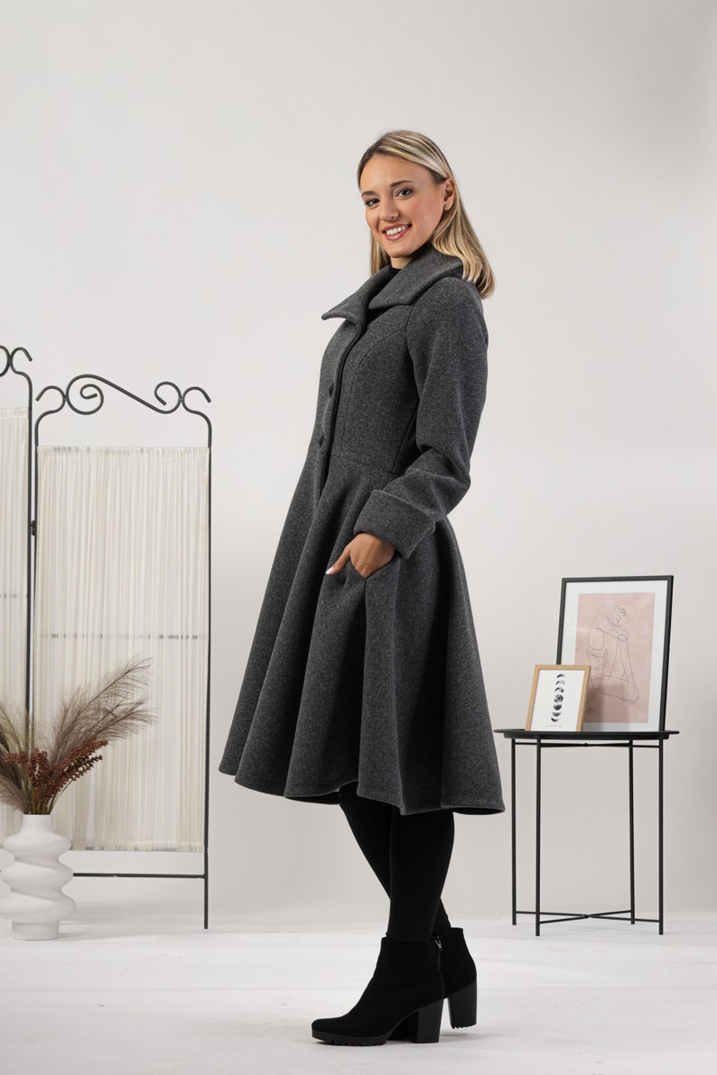 May include: A charcoal grey wool-blend coat featuring a wide collar, long sleeves with cuffs, a fitted bodice, and a flared, knee-length skirt with side pockets. The coat is fastened with a vertical row of buttons down the front centre.