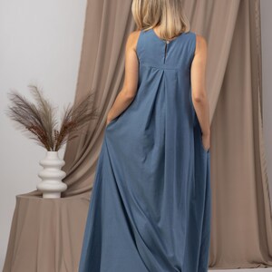 K&ouml;nnte beinhalten: Ein &auml;rmelloses, bodenlanges blaues Kleid mit Knopfverschluss am R&uuml;cken. Das Kleid hat ein plissiertes Design am Oberteil und Taschen. Das Model steht vor einem neutralfarbenen Vorhang.