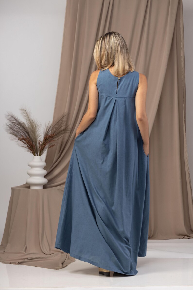K&ouml;nnte beinhalten: Ein &auml;rmelloses, bodenlanges blaues Kleid mit Knopfverschluss am R&uuml;cken. Das Kleid hat ein plissiertes Design am Oberteil und Taschen. Das Model steht vor einem neutralfarbenen Vorhang.