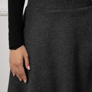 May include: A dark gray flared skirt with a wide waistband. The skirt is made of a textured fabric and is paired with a black long-sleeved top. The image shows a close-up of the skirt's fabric and design.