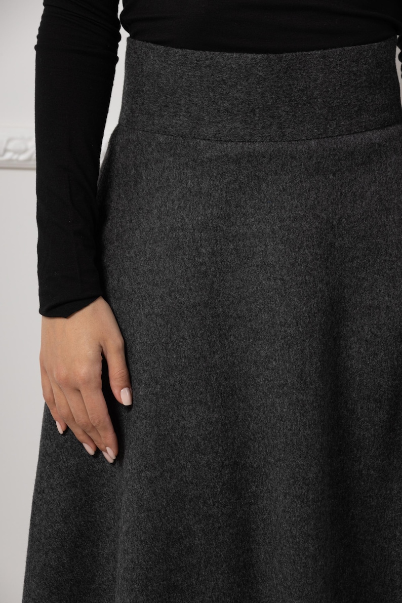May include: A dark gray flared skirt with a wide waistband. The skirt is made of a textured fabric and is paired with a black long-sleeved top. The image shows a close-up of the skirt's fabric and design.