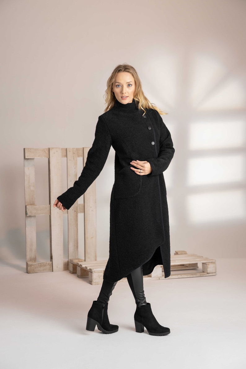 May include: A long black coat with a high collar and asymmetrical hemline. The coat features three large buttons and two pockets. The model is wearing black boots and leggings. The coat is made of a textured fabric.