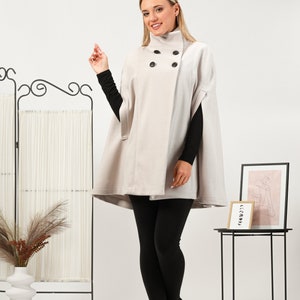 May include: A beige double-breasted cape coat featuring a high collar, four black buttons, and arm slits. The garment is worn over a black long-sleeved top, black leggings, and black ankle boots, creating a layered look with a structured silhouette.