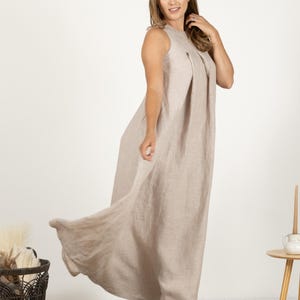 K&ouml;nnte beinhalten: Ein &auml;rmelloses, bodenlanges beiges Leinenkleid. Das Kleid hat ein plissiertes Oberteil und einen flie&szlig;enden Rock. Das Model tr&auml;gt beige Schuhe. Ein Korb und ein kleiner Tisch sind im Hintergrund.