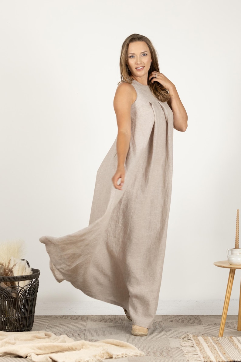 K&ouml;nnte beinhalten: Ein &auml;rmelloses, bodenlanges beiges Leinenkleid. Das Kleid hat ein plissiertes Oberteil und einen flie&szlig;enden Rock. Das Model tr&auml;gt beige Schuhe. Ein Korb und ein kleiner Tisch sind im Hintergrund.