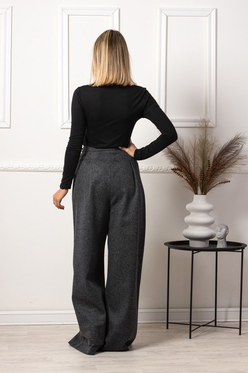 May include: A black long-sleeve top paired with high-waisted, wide-leg gray trousers. The pants have a tailored look. The outfit is displayed in a well-lit room with a decorative vase and side table.