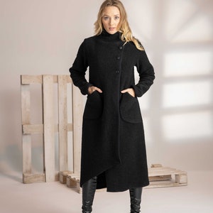 May include: A long black coat with a high collar and asymmetrical hem. The coat features large pockets and a button closure. The fabric appears to be a textured wool blend. The model is wearing black boots.