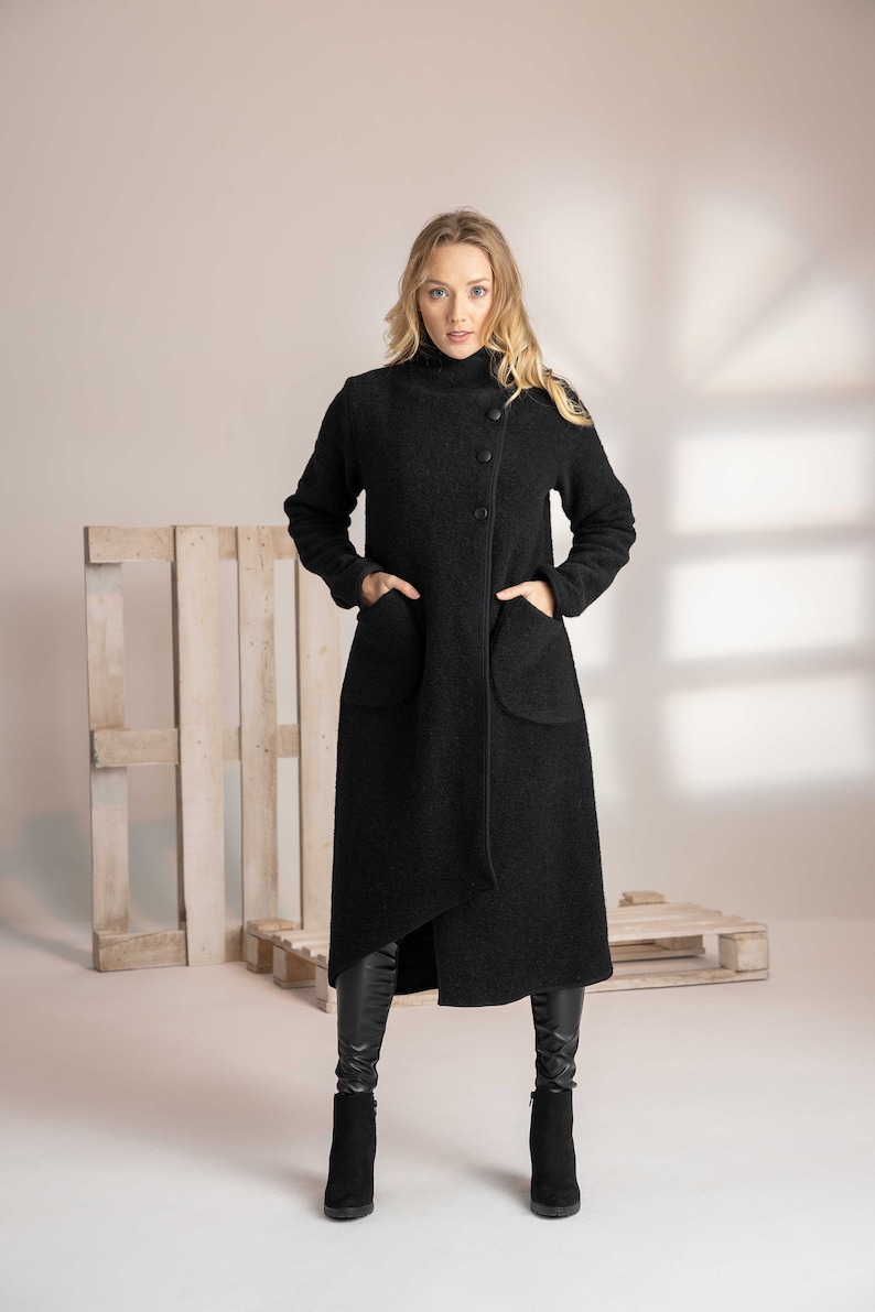May include: A long black coat with a high collar and asymmetrical hem. The coat features large pockets and a button closure. The fabric appears to be a textured wool blend. The model is wearing black boots.