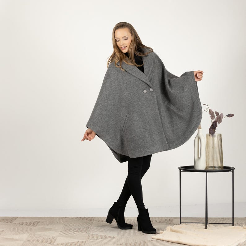 May include: A gray cape with a double-breasted button closure and two front pockets. The cape is worn over a black top and black leggings. The model is wearing black heeled boots. A small black side table with a vase and bottle is in the background.