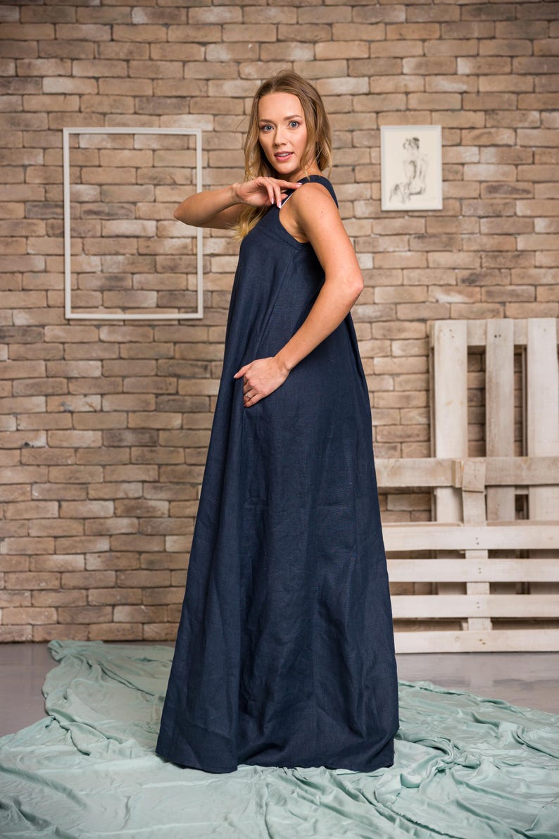 K&ouml;nnte beinhalten: Ein &auml;rmelloses, marineblaues Maxikleid mit flie&szlig;ender Silhouette. Das Kleid hat einen runden Ausschnitt und Taschen. Das Model steht vor einer Backsteinmauer.