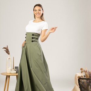 Może przedstawiać: Oliwkowa sp&oacute;dnica z gorsetowym stanikiem z czarnymi guzikami i sznurowaniem. Sp&oacute;dnica ma pełny, zwiewny kr&oacute;j. Modelka ma na sobie biały t-shirt. Sp&oacute;dnica ma kieszenie.